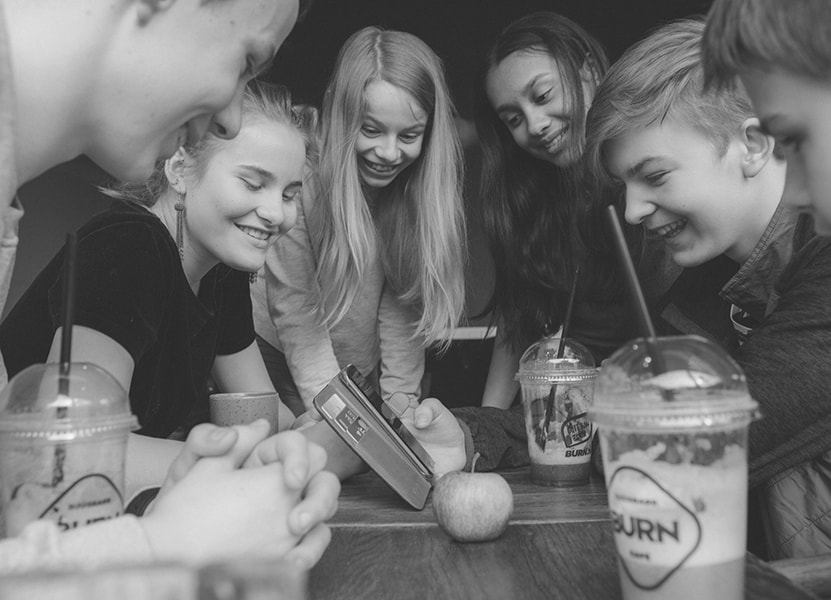 Sort/hvidt billede at fem teenagere rundt om en mobil på en café